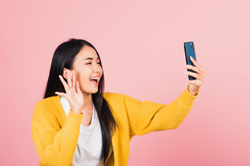 Wall Mural - Happy Asian portrait beautiful cute young woman smiling excited  making selfie photo, video call on smartphone studio shot isolated on pink background, female hold mobile phone raise hand say hello