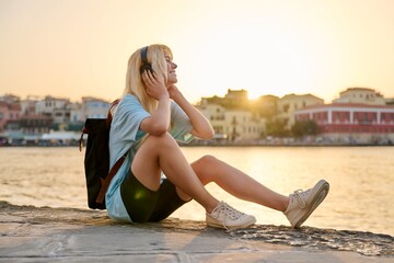 Wall Mural - Happy teenage female tourist sitting on sea promenade of an old city in sunset