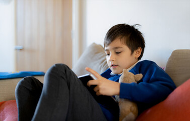 Wall Mural - Happy schoolboy playing with dog toy and game online with friend on tablet,Kid using internet sending homework to the teacher, Positive child sitting on sofa relaxing in living roomafter school