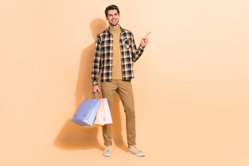 Poster - Full length body size view of attractive cheery guy carrying bags demonstrating offer isolated over beige pastel color background