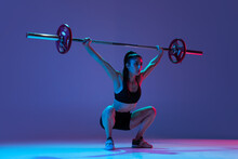 Portrait Of Muscled Woman In Sportswear Training With A Barbell Isolated On Purple Background In Neon Light. Sport, Weightlifting Concept