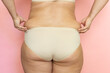 © Юля Бурмистрова - Woman with thick legs and buttocks closeup, clutch sagging folds on back, fat and cellulite. Naked overweight plus size girl on pink background in beige underwear. Concept of dieting and body control.