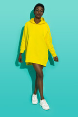 Wall Mural - Young black woman is posing in vibrant yellow oversized hooded sweatshirt and white sneakers.
