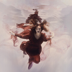 Wall Mural - A woman in a red airy dress swims under water on a light background as if flying