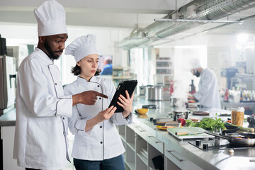 Wall Mural - Food industry workers with handheld touchscreen device preparing ingredients for evening dinner service at restaurant. Gourmet cuisine experts with modern tablet researching ingredients on internet.