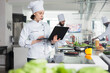 © DC Studio - Sous chef with modern laptop looking for gourmet dish recipe while standing in restaurant professional kitchen. Food industry worker with handheld computer brainstorming garnish ideas.