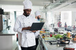© DC Studio - African american head chef with laptop searching for dish recipes on internet while standing in restaurant kitchen. Professional cook wearing cooking uniform using computer to inspire for gourmet meal