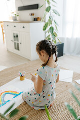 Wall Mural - A happy girl draws rainbow sitting on carpet floor in living room. Arts and crafts.