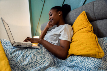 Wall Mural - Young African American woman laying in bed in the bedroom using laptop.