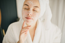 Young Beautiful Woman Wearing Bathrobe And Towel On Her Hair Applying Moistrizing Cream On Her Face. Skin Care Morning Rituals. Beauty Routine.
