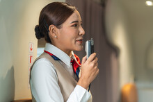 Asian Female Flight Attendant Holds A Microphone Talking To Passengers On Board An Airliner Serving Passengers During Their Journey.