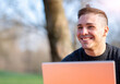 © Massimo Parisi - Portrait of attractive young office worker in casual wear, smiling in a break moment. He works remotely with the laptop from a table in the park, remote working concept