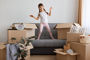 Indoor shot of happy smiling little girl wearing casual style attire standing on sofa and dancing, being happy to move into a new apartment, expressing positive emotions.