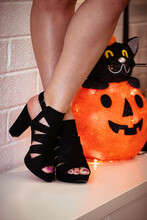 Feet In Black Heels Standing Crossed In Front Of A Paper Mache Jack-o-lantern With A Cartoon Cat