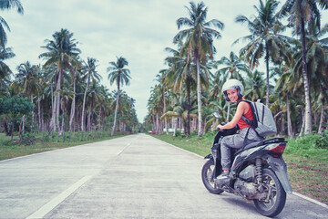 Canvas Print - Tropical travel and transport. Young beautiful woman in helmet riding scooter on the road with palm trees.