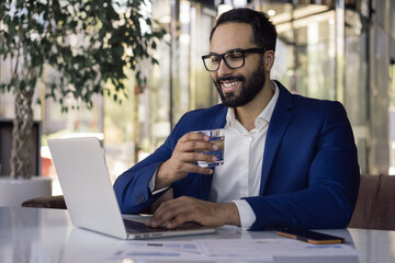 Wall Mural - Handsome smiling Moroccan businessman using laptop computer working online sitting in modern office. Portrait of happy successful manager drinking water planning project sitting at workplace