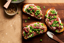 A Pea, Prosciutto, And Ricotta Crostini With A Bite Missing On A Plate Next To A Bowl Of Peas, Walnuts, And Flakey Sea Salt.
