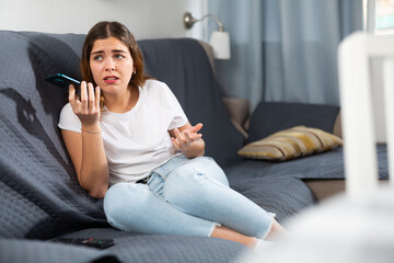 Dissatisfied young woman complaining to anyone on mobile phone while sitting on sofa at home