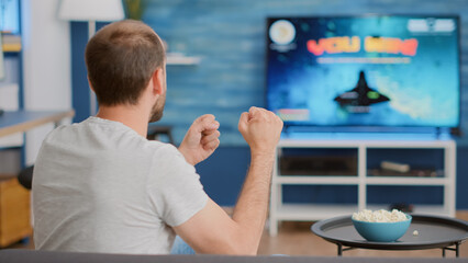 Sticker - Closeup of man holding controller celebrating victory in online first person shooter game in front of big screen tv. Gamer doing celebration hand gesture happy with performance in gaming simulation.