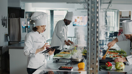 Wall Mural - Authentic chef looking at food recipe on digital tablet screen, using online internet website to make gastronomy dish. Woman cook with device making professional cuisine restaurant meal.