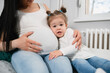 © Dragica - Amazing beautiful little girl with ponytails cuddles with her pregnant mom on mother's belly.Caucasian pregnant female fondles her tummy with her right hand and hugs her daughter with her left hand.