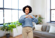 people, real estate and mortgage concept - happy smiling woman in blue sweater and jeans showing hand heart gesture over home living room background