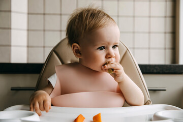 10 months old baby sitting in high chair, with a silicone bib, eating finger food.