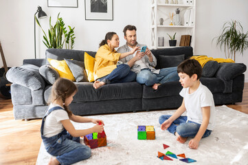 Two parents using smartphones sitting on the sofa in cozy living room, scrolling news feed while two kids playing with constructor on the floor, sibling enjoying game together. Family of four at home