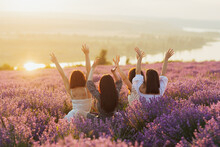 Beautiful Girlfriends Having Picnic In The Lavender Field In Summer Sunset. They Sitting With Hands Up And Enjoying Beautiful Landscape. 