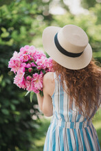 Portrait Of Young Redhead Curly Woman In Straw Hat And Linen Stripe Dress With A Basket And A Pink  Peonies Bouquet In The Garden. Back View