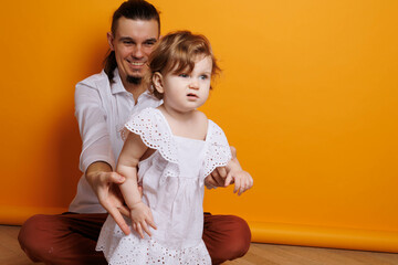 Little baby girl standing on white chair with yellow background. Serious long-haired little one-year daughter sitting on chair alone. Blonde-haires girl surprised. Sweet little girl smiling