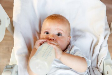 Cute little newborn girl drinking milk from bottle and looking at camera on white background. Infant baby sucking eating milk nutrition lying down on crib bed at home. Motherhood happy child concept