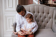 © Tatyana - Handsome happy father sitting with his little daughter on beige sofa indoors. Young man in white shirt with his 1-year-old daughter in white dress, holding her in hands on legs. family tenderness