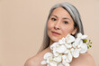 Beautiful romantic caucasian mature middle-aged woman with grey hair looking at camera covering her face with orchid flower isolated in beige background. Beauty and skin hair care concept