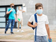 Leinwandbild Motiv Young boy in face mask standing at entrance to school building. Senior students walking in background.