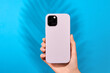 Female hand holding white smartphone in soft silicone cover back view . Phone case mock up isolated on blue background shadow from a plant