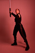 © faestock - Full length portrait of pretty redhead female model wearing black futuristic scifi leather cloak costume. Standing pose  holding lightsaber on red studio background with coloured lighting