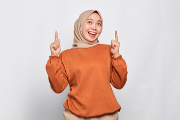 Wall Mural - Amazed young Asian woman in orange shirt pointing finger up at copy space isolated over white background
