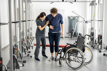 Rehabilitation Specialist Helps A Guy Stand Out Of A Wheelchair At Rehabilitation Center. Concept Of Physical Therapy And Support For People With Disabilities