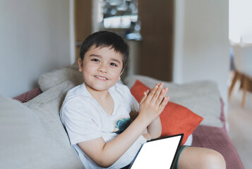 Wall Mural - Happy young boy looking at camera with smiling face and clapping hands, Positive kid using tablet playing game online at home, Child sitting on sofa relaxing on weekend