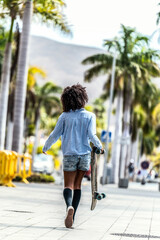 Wall Mural - Back view of the walking girl with afro hairstyle, carrying skateboard in hand. Summer vibes. Hobby concept