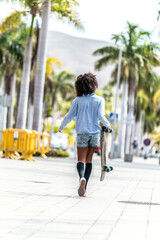 Wall Mural - Back view of the walking girl with afro hairstyle, carrying skateboard in hand. Summer vibes. Hobby concept