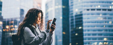 Young Woman Holds Smartphone And Flips Through Information On Gadget Screen Standing Against Big Modern Financial Centers In Evening
