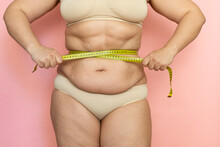 Cropped Photo Of Naked After Birth Woman Tummy In Underwear, Trying To Lose Weight. Holding And Measuring Her Waistline By Roulette Tape Belly, Abdomen. Fat Large Sagging Folds On Stomach. Excess Skin
