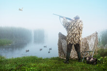 Hunter Shooting Into Sky During Duck Hunting At Sunrise. Hunting With Ducks Decoy On River Bank.