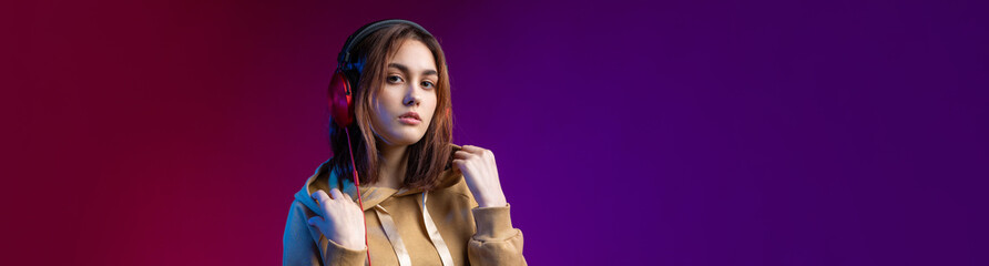 Wall Mural - Young beautiful fashionable hipster girl dressed in a hoodie  listening to music in red headphones in a studio red dramatic background