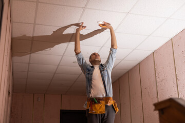 Handsome young builder is twisting the light bulb in. The man is looking up.