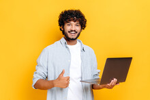 Positive Attractive Arabian Or Indian Guy With Curly Hair, Wearing Casual Clothes, Holding An Open Laptop In His Hand, Looks At Camera, Showing Thumbs Up Gesture, Stands On Isolated Orange Background