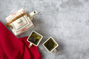 A Chinese-style square-shaped porcelain teapot and two tea mugs and a red towel are on a gray background. Top view or flat lay. Copy space for text or design. Theme of International Tea Day.