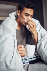 Wall Mural - Dont get the flu, get the shot. Shot of a young man blowing his nose and having tea while recovering from an illness in bed at home.